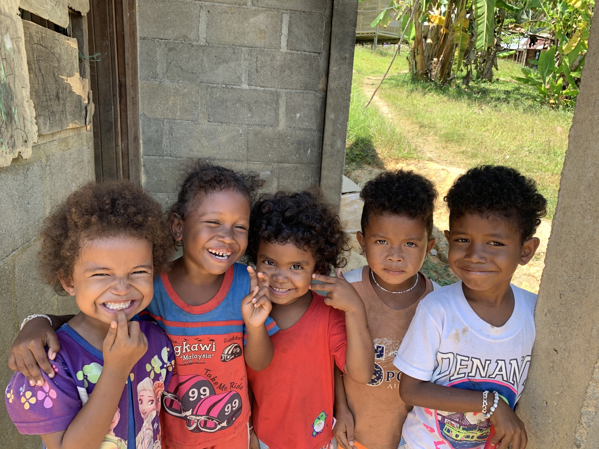 Five smiling Orang Asli children standing together in their village, representing the communities Saora+ serves